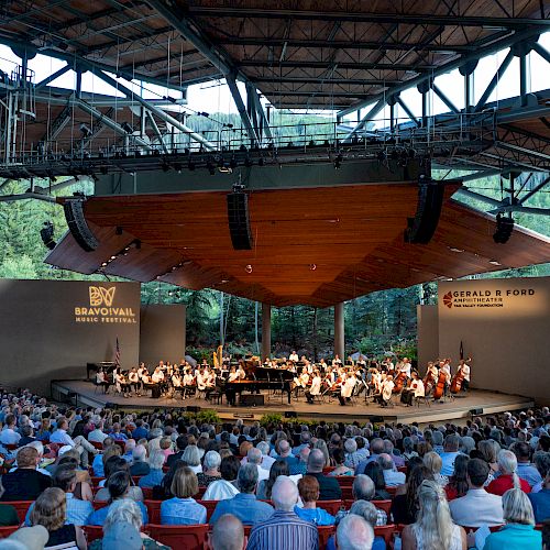 An outdoor concert with a symphony orchestra performing on stage, surrounded by a large audience under a wooden canopy structure in a forested area.