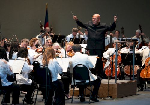 An orchestra performs on stage, with a conductor leading them. Musicians play various instruments, including strings and brass, in a concert setting.