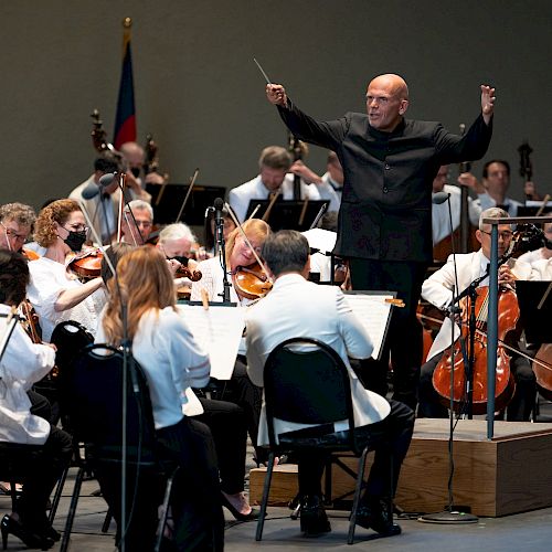 An orchestra performs on stage, with a conductor leading them. Musicians play various instruments, including strings and brass, in a concert setting.