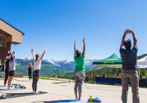 A group of people is doing yoga outside on mats with a scenic mountain view and clear blue skies, participating in a standing stretch.