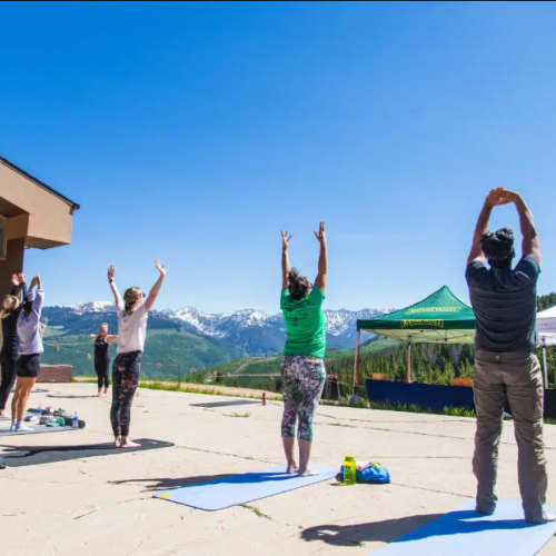 A group of people is doing yoga outside on mats with a scenic mountain view and clear blue skies, participating in a standing stretch.